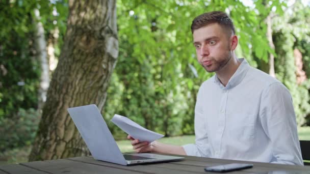 Молодой человек читает и пользуется ноутбуком в парке — стоковое видео