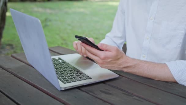Manos de hombre usando un ordenador portátil y un teléfono en el parque — Vídeos de Stock
