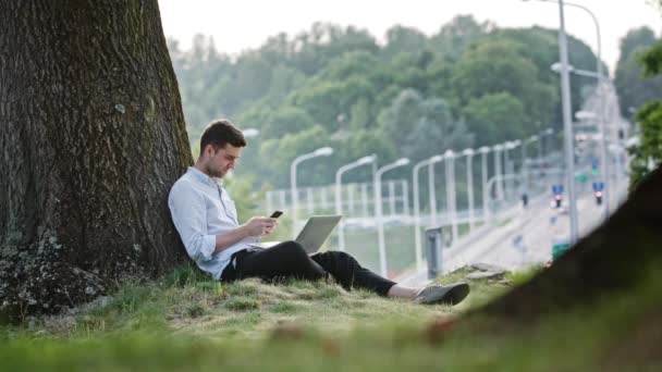 Un joven usando un móvil y un ordenador portátil al aire libre — Vídeos de Stock