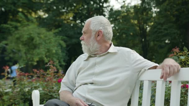 Старик сидит на белой скамейке в парке — стоковое видео