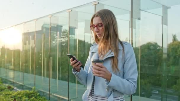 Een jonge dame lopen en met behulp van een telefoon buitenshuis — Stockvideo