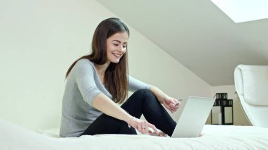 Bir yatağa evde oturan bir dizüstü bilgisayar kullanan kadın