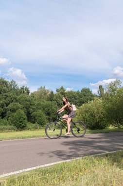 Kız bir bisiklet sürme. Yan görünümü. Orman ve arka planda bulutlar