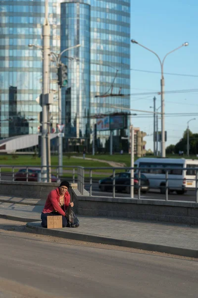 Бездомный сидит на тротуаре с картонкой и надписью: нужна работа. На заднем плане бизнес-центр — стоковое фото