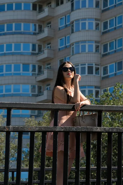Modern bir kız elbise, gözlük ve bir çanta bir köprü büyük bir bina arka plan üzerinde duruyor. Modern genç iş kadını