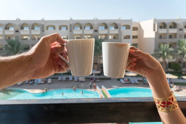 男性和女性的手保持在阳台上的咖啡杯背景下的酒店，建筑和游泳池都可见 — 图库照片