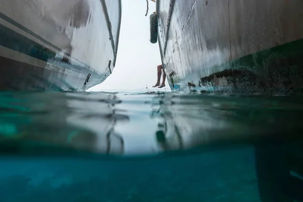 I havet finns det två båtar och från sidan syns benen av en man. Visa hälften under vatten — Stockfoto