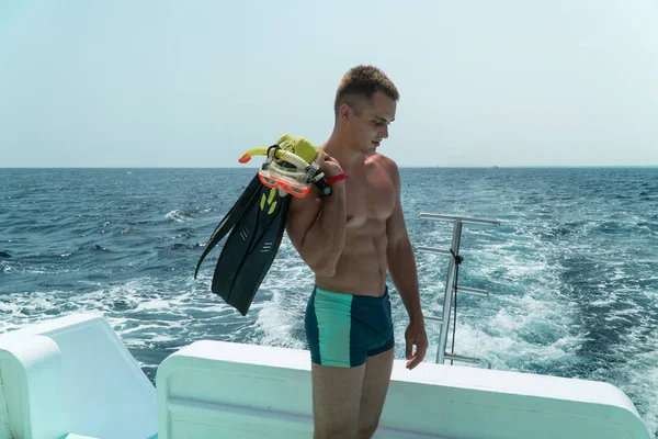 The guy stands in the slats in the stern of the yacht with fins and a mask in his hands. The guy is preparing to dive under water