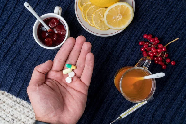 Bir adamın elini bir hap tutar. Çay limon, tabletler ve termometre, reçel ve çilek ile natürmort. Soğuk algınlığı tedavisinde kavramı