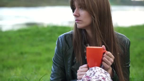 Una chica está bebiendo té caliente mientras está sentada en la hierba cerca del río . — Vídeo de stock