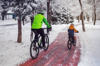 Bir adam ve oğlu bir kış parkında bisiklet yolu boyunca bisiklet sürerler.