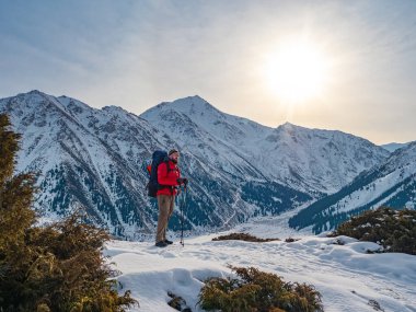 A tourist in a bright red jacket stands against the backdrop of a magnificent mountain landscape on a sunny morning