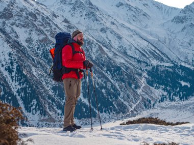 A tourist in a bright red jacket stands against the backdrop of a magnificent mountain landscape on a sunny morning