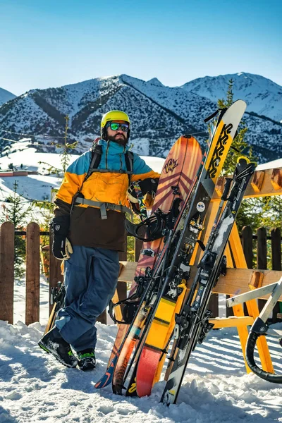 Un tipo barbudo con casco y gafas se encuentra cerca de un estante con esquís y tablas de snowboard. Hermoso día soleado en la estación de esquí —  Fotos de Stock