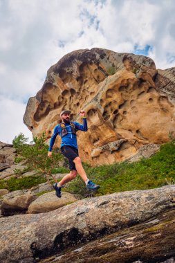 Adam dağ yolunda koşuyor. Dağlarda, kayaların arasında koşan patika. Mavi formalı ve siyah şortlu bir adam dışarıda antrenman yapıyor.