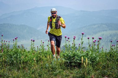 Atletik adam çayırların arasında dağlarda koşuyor. Sarı tişörtlü ve siyah şortlu bir adam açık havada antrenman yapıyor. İz çalışıyor