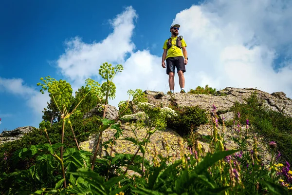 Athlete runner stands on the edge of a cliff in the mountains. Man in a yellow T-shirt and black shorts is training in outdoors. Trail running