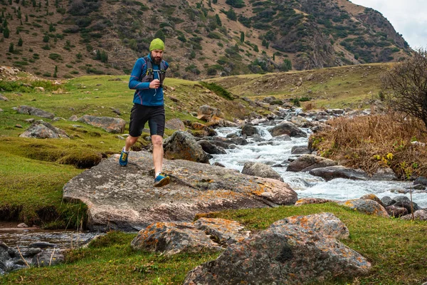 Runner guy is jogging in the highlands. Athlete runs near a mountain river. Man is training outdoors. Trail running