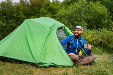 Elinde metal bir kupayla yeşil bir çadırın yanında oturan neşeli bir turist. Yazın seyahat ediyorum.