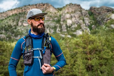 Kayalardan oluşan bir arka planda koşan bir sporcunun portresi. Mavi kazaklı ve koşu yelekli bir adam. İz çalışıyor