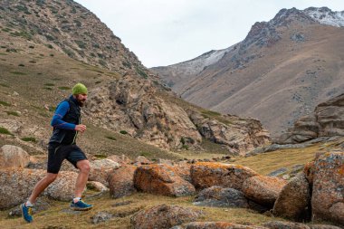 Sakallı bir sporcu dağlarda dışarıda koşuyor. Dağların zirvesinde antrenman yapıyor. Gökyüzü çalışıyor