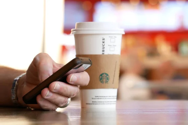 Seorang pria membaca pesan di ponselnya dan meminum kopi panas Starbucks dengan orang-orang gerakan kabur di area food court — Stok Foto