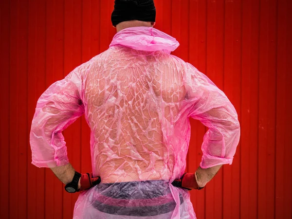 Un homme se tient sous la pluie dos dans le chapeau noir, gants, un dozhdivike en plastique rose transparent avec une capuche sur un fond rouge Photos De Stock Libres De Droits