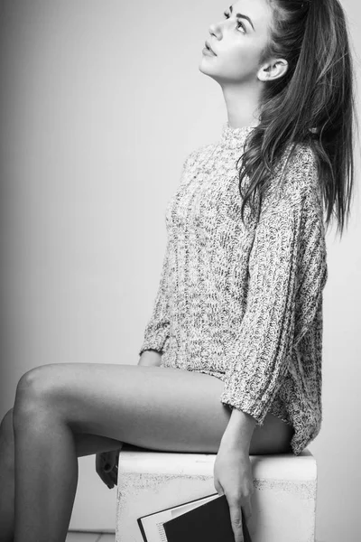 Woman holding a book. Sensual brunette woman with long hair in gray pullover sitting on white cube over light grey studio background The beautiful young girl. Sexual forms. Burning person. High slender figure. Stately image. 