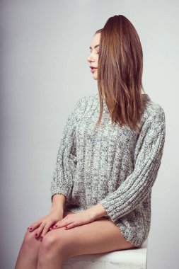 Genç bir kız bir küp üzerinde oturur. Çıplak dizlerinin üzerinde ellerini koymak. Bir gri kazak. Fotoğraf stüdyosu içinde photoshoot.