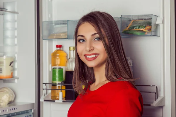 Vrouw rode jurk koelkast deur, bereiken binnen kijken naar je camera glimlachen — Stockfoto