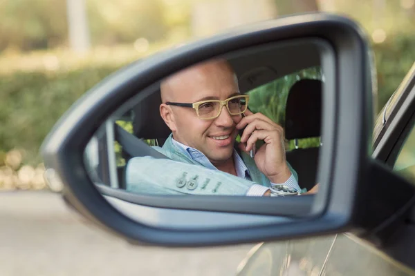 Guapo hombre de negocios feliz en el teléfono móvil en el coche. Reflexión en el espejo lateral — Foto de Stock