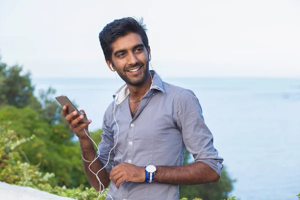 Retrato de primer plano, joven con camisa azul y gafas negras, escuchando auriculares, viendo canciones en su teléfono, fondo aislado al aire libre — Foto de Stock