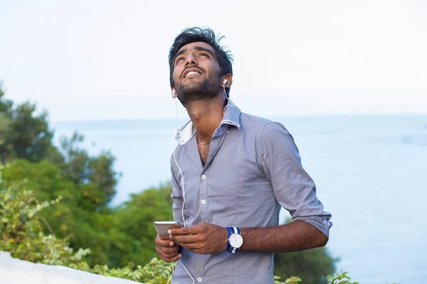 Primo piano Ritratto di un bel ragazzo felice in camicia blu guardando verso l'alto nel pensiero ascoltando musica, rilassandosi all'aperto, isolata verde alberi fondo mare . — Foto Stock