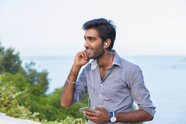 Retrato de primer plano, joven con camisa azul y gafas negras, escuchando auriculares, viendo canciones en su teléfono, fondo aislado al aire libre — Foto de Stock