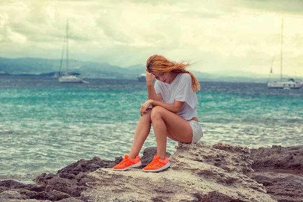 Triste joven mujer seria sentada en la roca mirando hacia abajo con el fondo del cielo marino — Foto de Stock