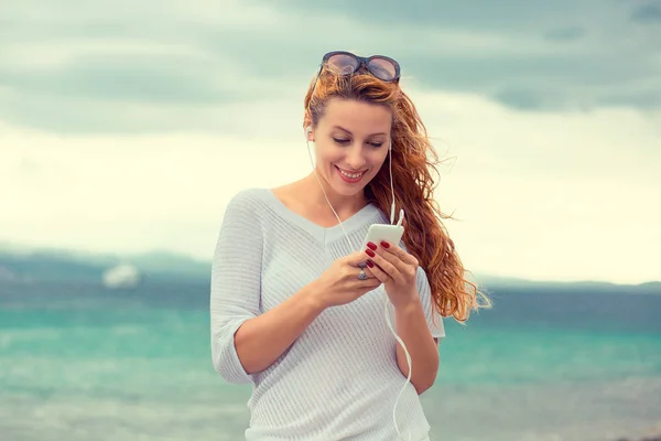 Femme heureuse en lunettes de soleil textant sur un téléphone intelligent sur la plage avec la mer en arrière-plan. Effet de filtre Instagram appliqué — Photo