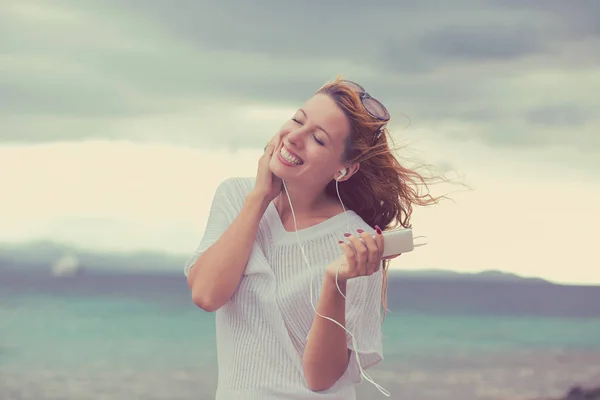 Potret seorang gadis muda yang tersenyum dengan earphone mendengarkan musik di luar rumah di pantai dengan latar belakang langit biru laut — Stok Foto