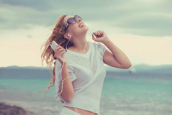 Portret van een lachende jong meisje met koptelefoon luisteren naar muziek buiten aan het strand met diepblauw hemelachtergrond — Stockfoto