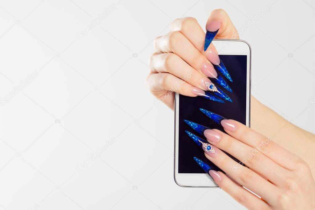 Woman hands with stiletto nails holding phone. Art Manicure. Modern style blue Nail Polish. Beauty hands holding mobile cellphone. Stylish Colorful stiletto Nails isolated white background
