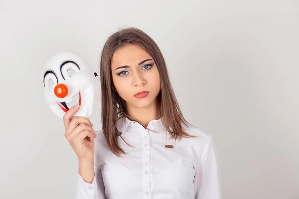 Retrato joven mujer preocupada disgustada con expresión triste sosteniendo una máscara de payaso expresando alegría felicidad aislada sobre fondo blanco de la pared. Sensación de expresión facial humana — Foto de Stock