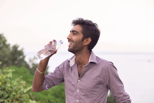 Idratati. Uomo sorridente di profilo in procinto di bere da una bottiglia di acqua alberi natura isolati e fondo mare — Foto Stock