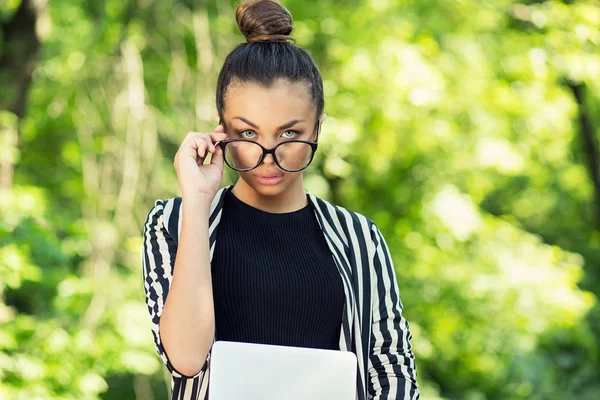 Skeptical. Doubtful Indian woman student looking at you camera holding eye glasses down holding laptop tablet computer isolated green park background — Stock Photo, Image