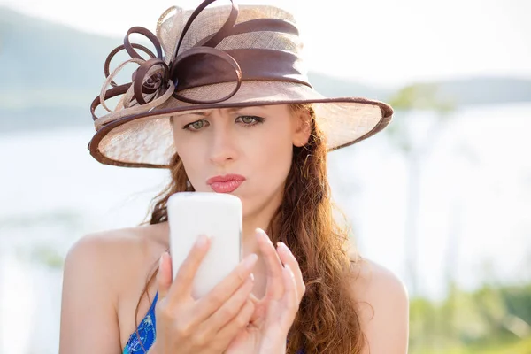 Portret de femeie tânără confuză în pălărie uitându-se la telefon, citind mesaje text neplăcute. Expresia feței reacției emoționale umane . — Fotografie, imagine de stoc