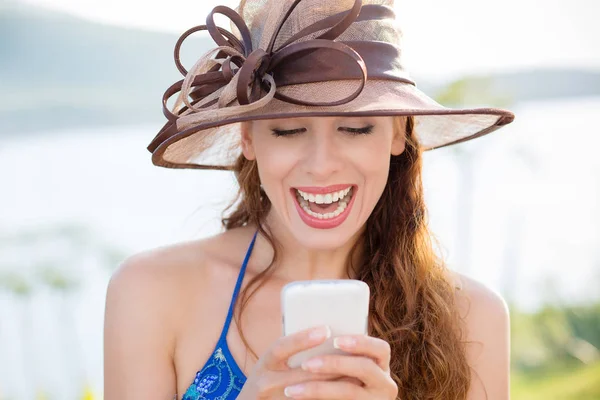 Closeup πορτρέτο ευτυχισμένη ενθουσιασμένος νεαρό κορίτσι στο καπέλο ψάχνετε στο τηλέφωνο με toothy χαμόγελο γέλιο βλέποντας αστείο ειδήσεις φωτογραφίες απομονωμένη φόντο λίμνη θάλασσα έξω πόλη. Θετικά συναισθήματα ανθρώπου, έκφραση του προσώπου — Φωτογραφία Αρχείου