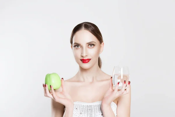 Привлекательная молодая женщина, смотрящая в камеру, держит в руках зеленое яблоко и стакан воды, пропагандирующие здоровый образ жизни, диету и спорт, изолированные на белом фоне. Лицо, язык тела . — стоковое фото