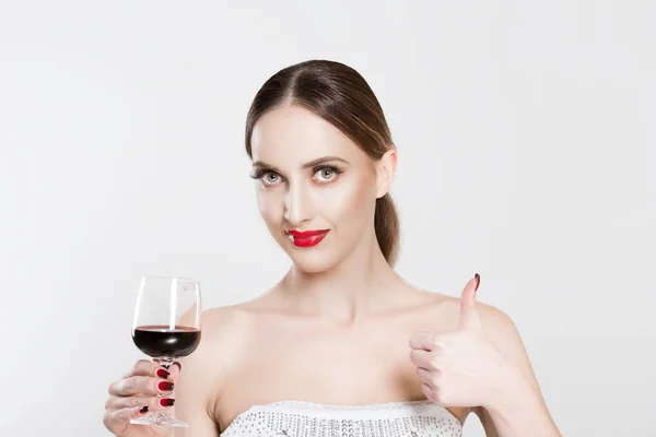 Портрет привлекательной молодой женщины, смотрящей в камеру, держащей бокал красного вина и показывающей большие пальцы вверх, изолированные на белом фоне стены. Позитивное человеческое выражение, жесты и язык тела . — стоковое фото
