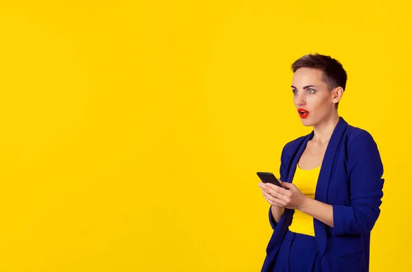 Mujer de negocios impactada con teléfono mirando al espacio de copia lateral aislado pared de fondo amarillo. Traje de negocios traje azul formal, pelo corto, labios rojos — Foto de Stock