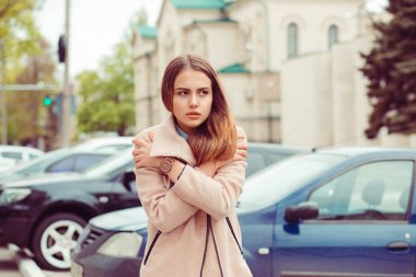 Güzel esmer kız giyinmiş bir bej bir palto almakla soğuk, kendini uzak seyir sarılma şehir araçları ve binaların arka plan üzerinde düşünme