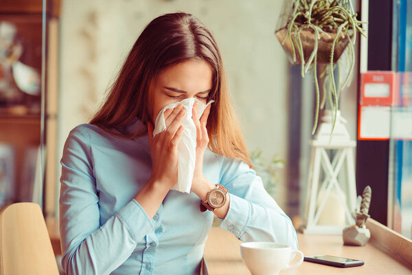Flu. Young woman got nose allergy, flu sneezing nose sitting at the table in a trendy cafe coffee shop with a cup of hot beverage beside