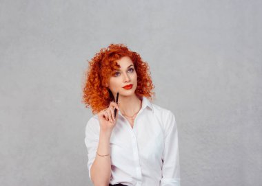 Şüpheli iş kadın düşünme kalem yanağına. Kırmızı kafa kıvırcık saç Bayan düşünceli kız beyaz resmi gömlek gri arka duvarda izole zencefil.